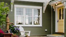 Front porch of a green house with a yellow door. A white chair, a faucet and some flowers are pictured in the foreground.
