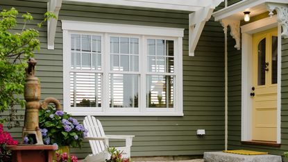 Front porch of a green house with a yellow door. A white chair, a faucet and some flowers are pictured in the foreground.