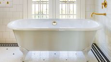 White roll-top freestanding bath with gold faucet on wall. The floor is tiled white with black geometric border. The walls having white subway tiles and there is a balcony door feature in the background