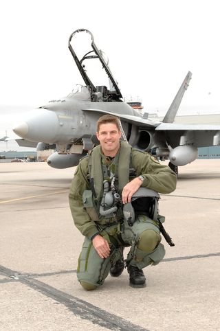 jeremy hansen kneeling on a runway in a flight suit. a fighter jet is parked a few feet behind him