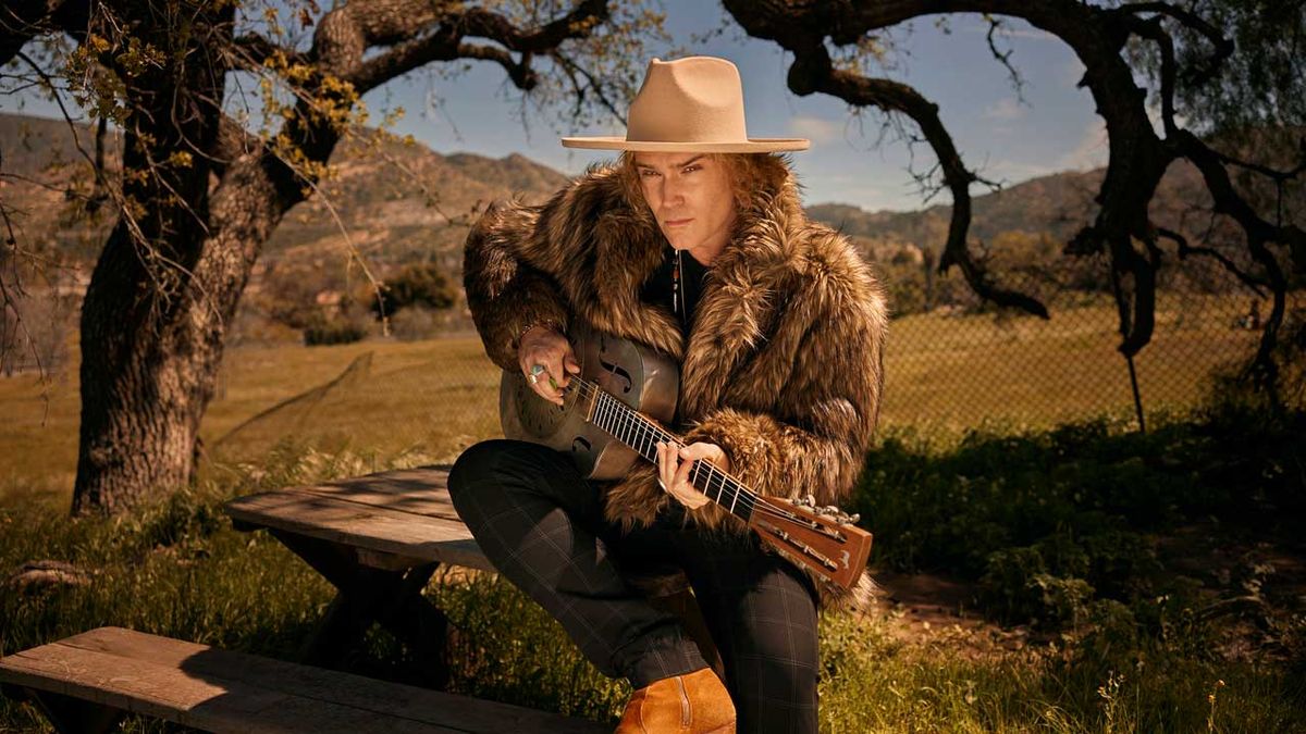 Philip Sayce sitting on a table beneath some trees holding an acoustic guitar