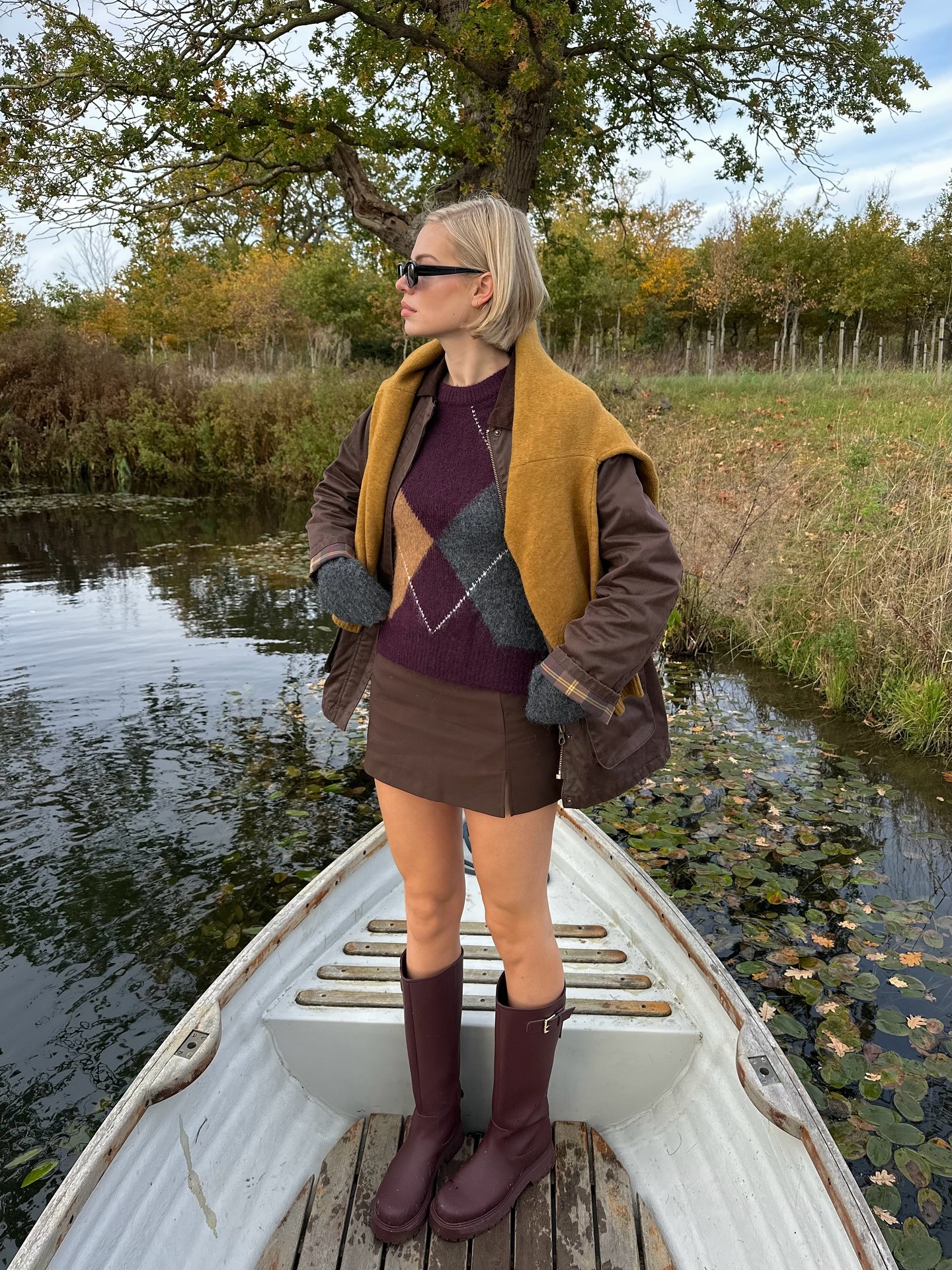 Fashion person Linda Sza wears winter knitwear trends 2025. She wears a argyle jumper with a brown mini skirt burgundy boots and a yellow jumper on her shoulders. This image was recently taken from her Instagram.