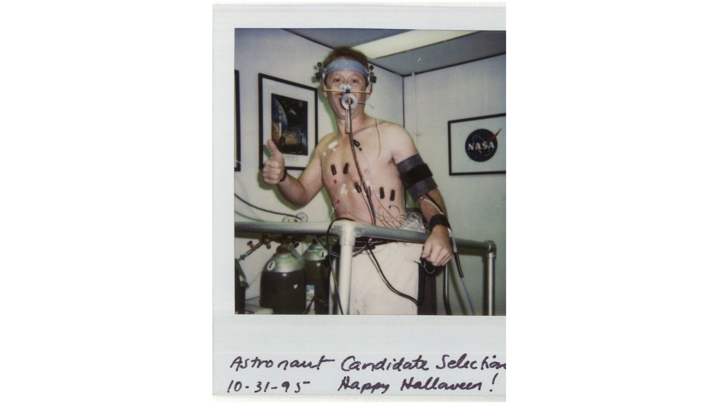 a shirtless man with electrodes attached to his chest gives a thumbs-up while on a treadmill in a white-walled room.