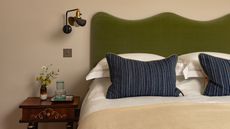 Bedroom with warm neutral walls, a green velvet headboard, striped navy pillows and a dark wood bedside table.