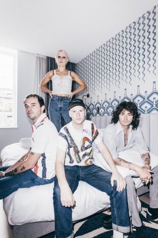 A portrait of Amyl And The Sniffers sitting on a hotel bed. Amy Taylor is hitting her bandmates with a pillow