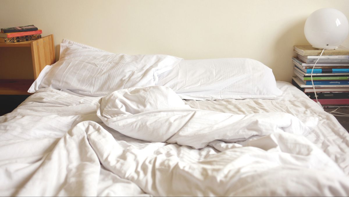 An unmade bed with a white duvet bunched up and white pillows, mattress shows slight body impressions