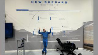 a man in a blue flight suit stands in front of a wall reading 