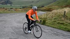 A cyclist in a orange jacket ride up a road with hills in the background