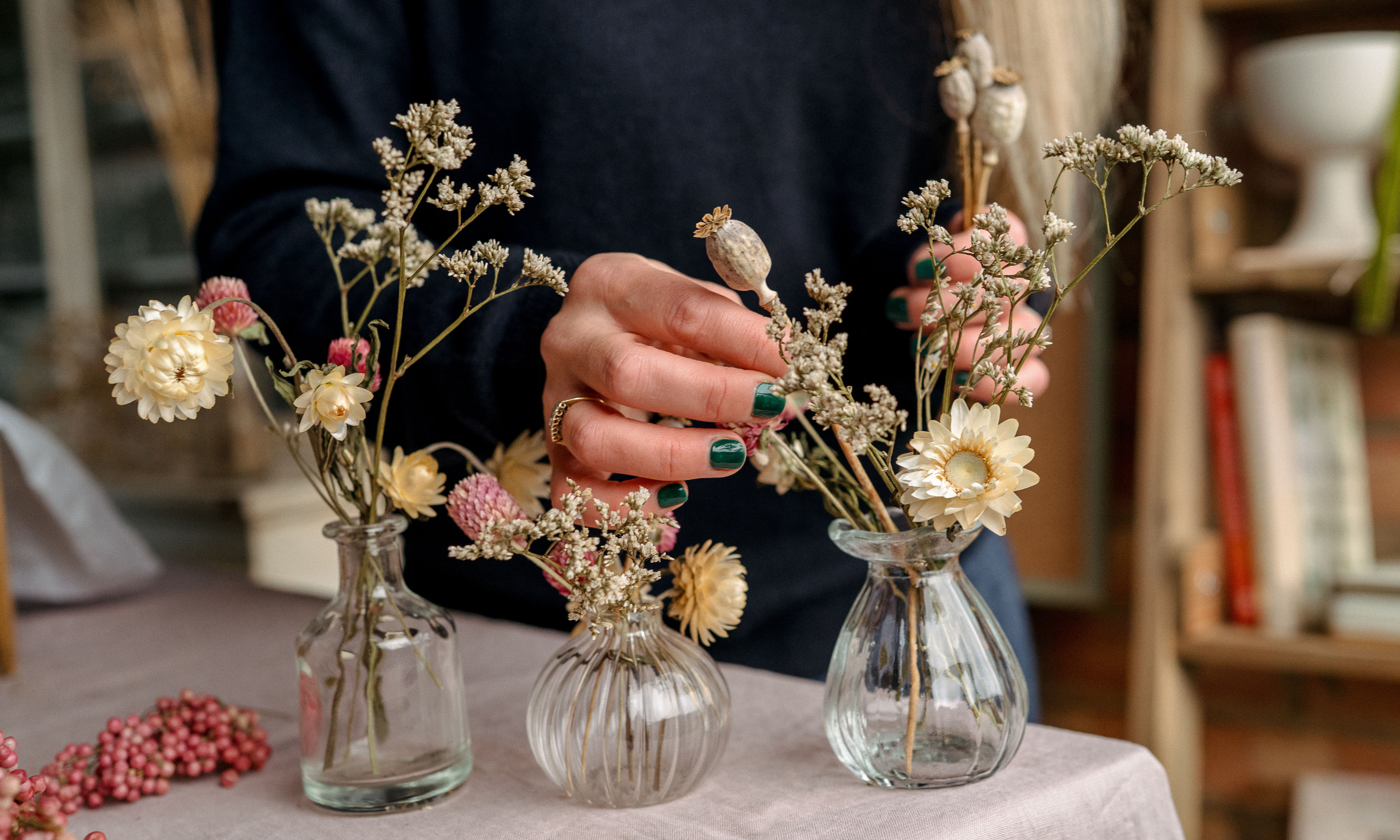 Hands putting dried flowers into three clear glass bud vases on a table
