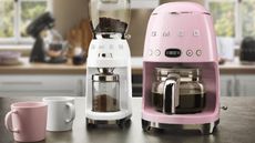 The pink smeg drip coffee maker next to a white smeg coffee grinder on a kitchen countertop with pink and white mugs