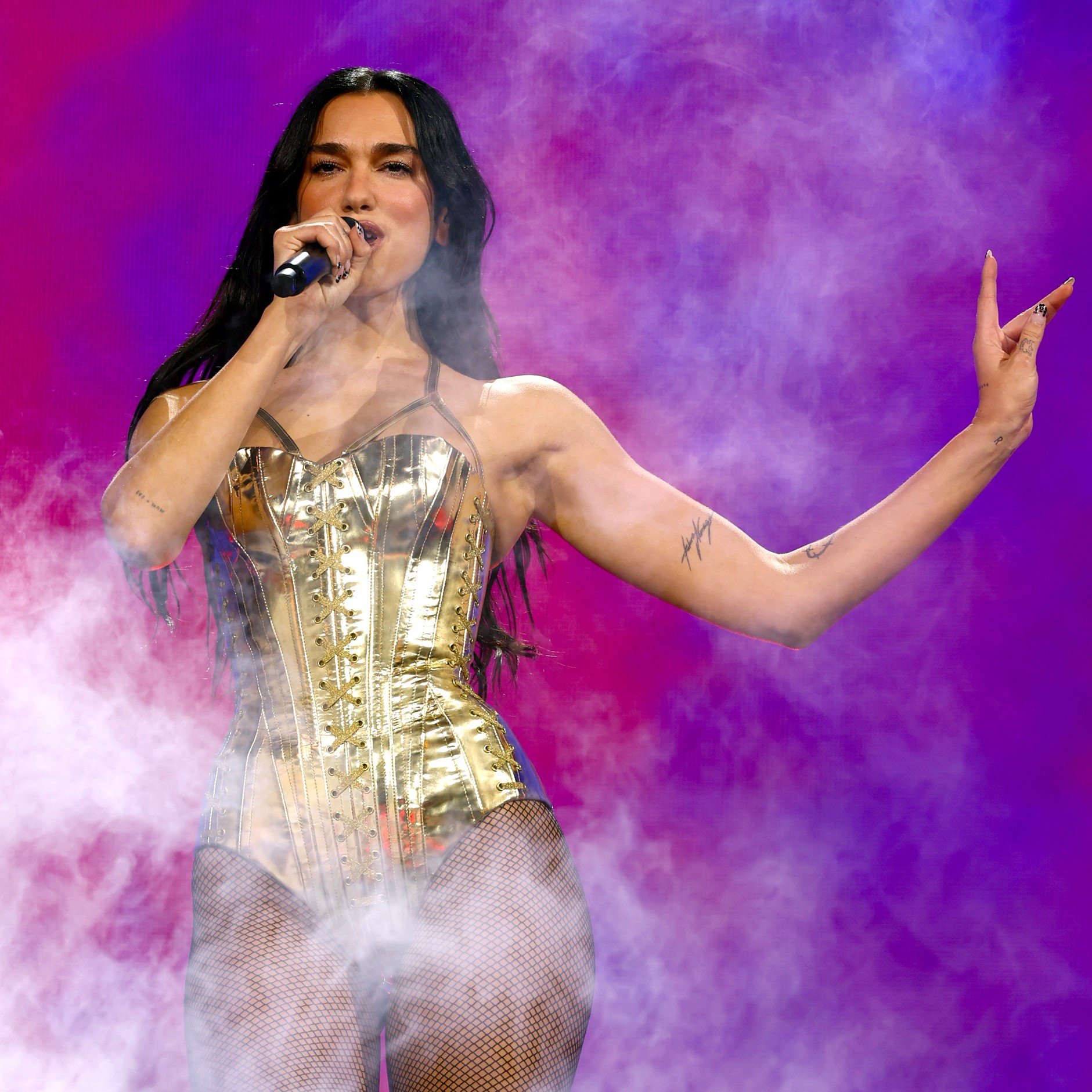 a photo of a woman with long dark hair wearing a gold leotard performing on stage in front of a pink and purple background