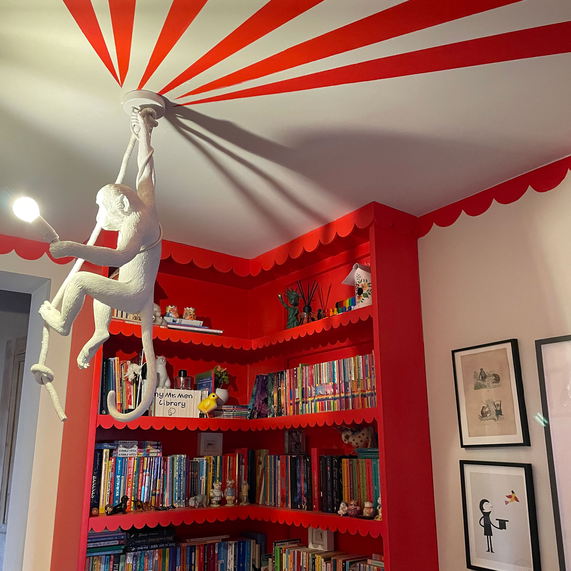Corner of room with red bookcase with scalloped details, white monkey lamp hanging from ceiling, with red stripes painted beside it