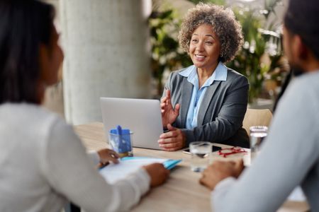 Happy mature financial adviser or insurance agent working on a computer while talking to her clients during a meeting in the office.