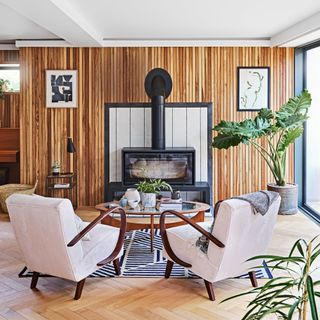 A modern traditional living room scheme with wood panel wall decor, two cream accent chairs and black wood-burning stove