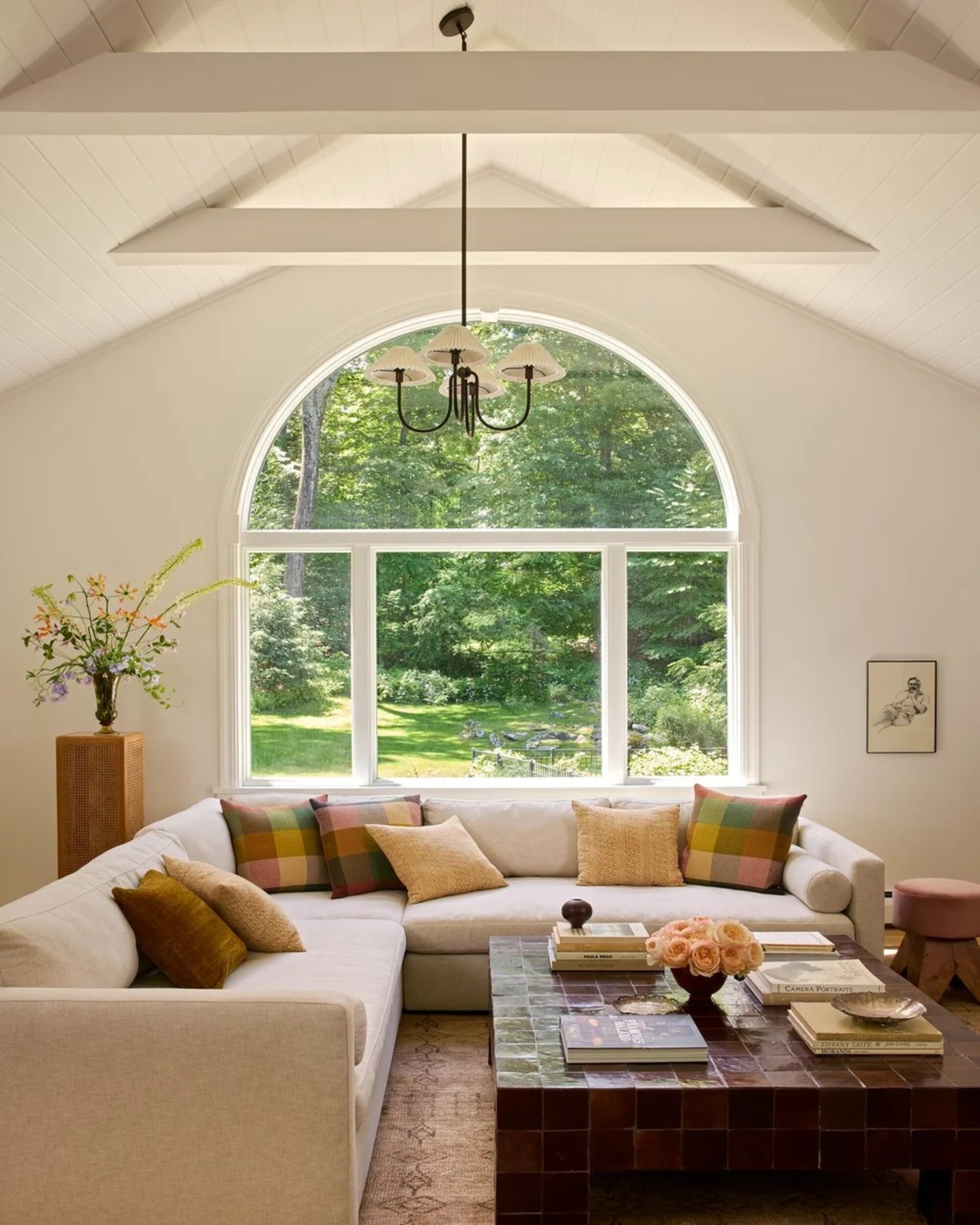 A white living room with an angled ceiling and a large, curved window on the back wall. There is a large off-white sectional sofa and a burgundy, tiled coffee table in the room.