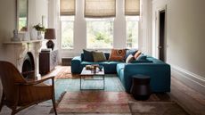 A neutral living room with a bright blue sofa, patterned cushions and patterned rugs. In the center of the room is a coffee table, and to the left is a large white mantelpiece with wooden chair. Behind are large windows with opaque beige blinds.