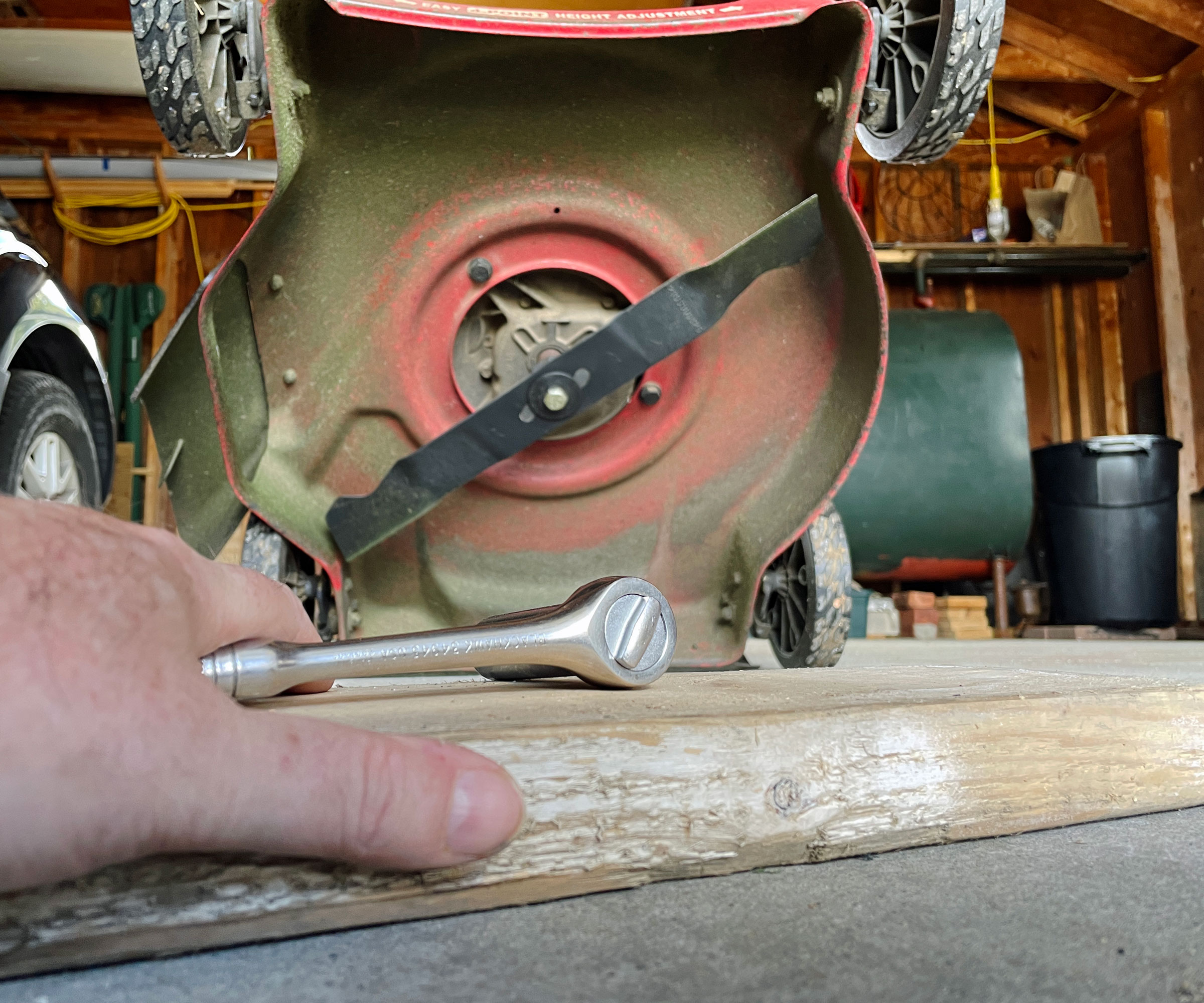 hand holding hand tool in front of lawn mower blade placed on wooden board