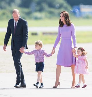 Prince William, wearing a suit, and Princess Kate, who is dressed in a purple long sleeved dress, holding hands with Prince George and Princess Charlotte walking on a sunny airport tarmac