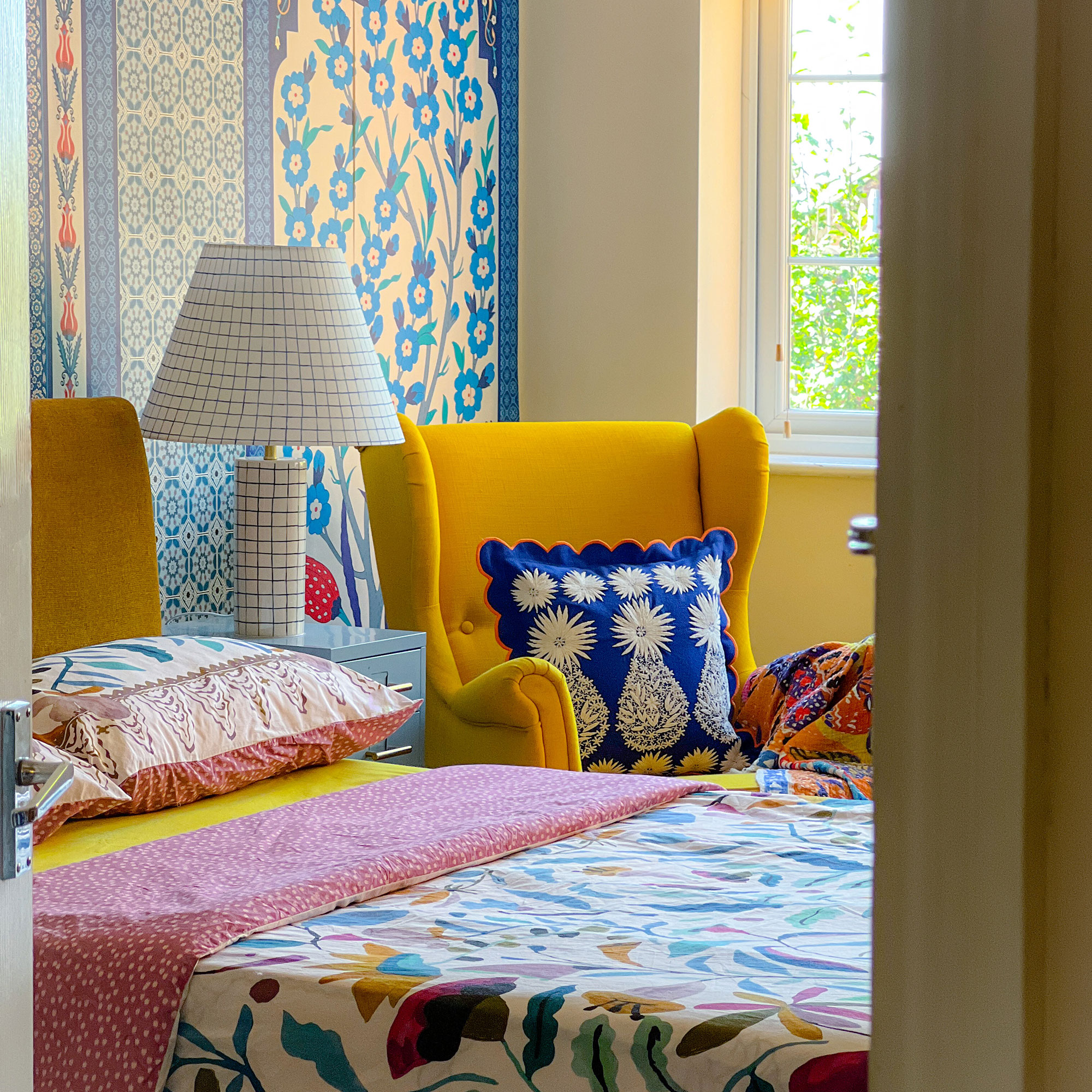 Bedroom with floral wall behind bed, floral duvet cover, checked bedside light and yellow chair with blue cushion