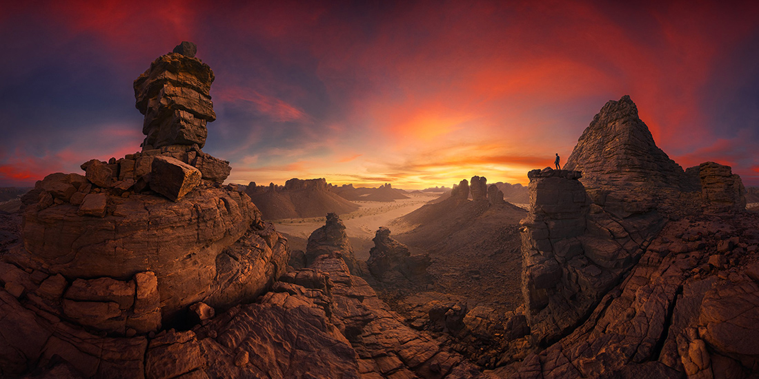 A vast desert landscape at sunset, featuring unique rock formations silhouetted against a vibrant sky with a lone figure on a peak