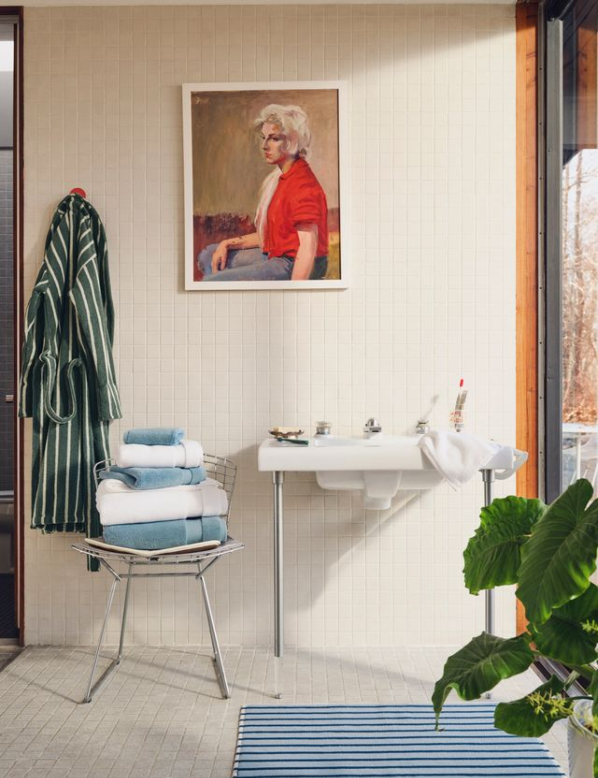 Retro-style bathroom covered in white ceramic tiles decorated with striped bath towels, a bath mat, and bath robe, alongside an oil portrait of a woman, which hangs overhead above the sink
