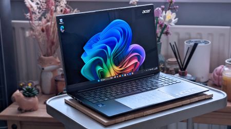 An Acer Swift Go 16 AI laptop sits on a small table, displaying the Windows 11 desktop. The keyboard and touchpad are visible, and the background includes decorative items like dried flowers.