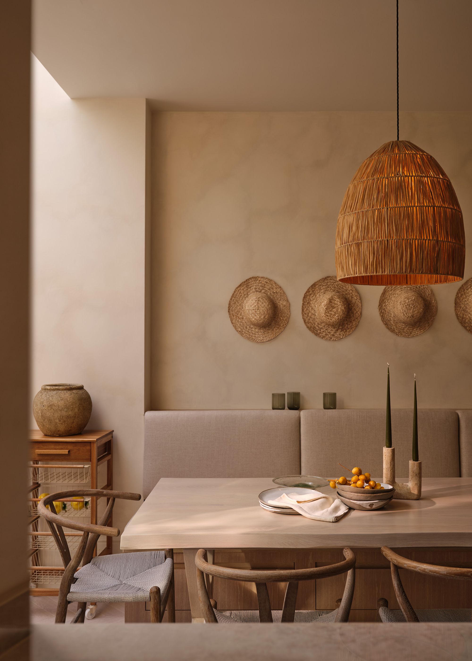 A dining room with plaster walls, a light wooden table with beige seating, beachy decor on the walls, and a woven pendant light.