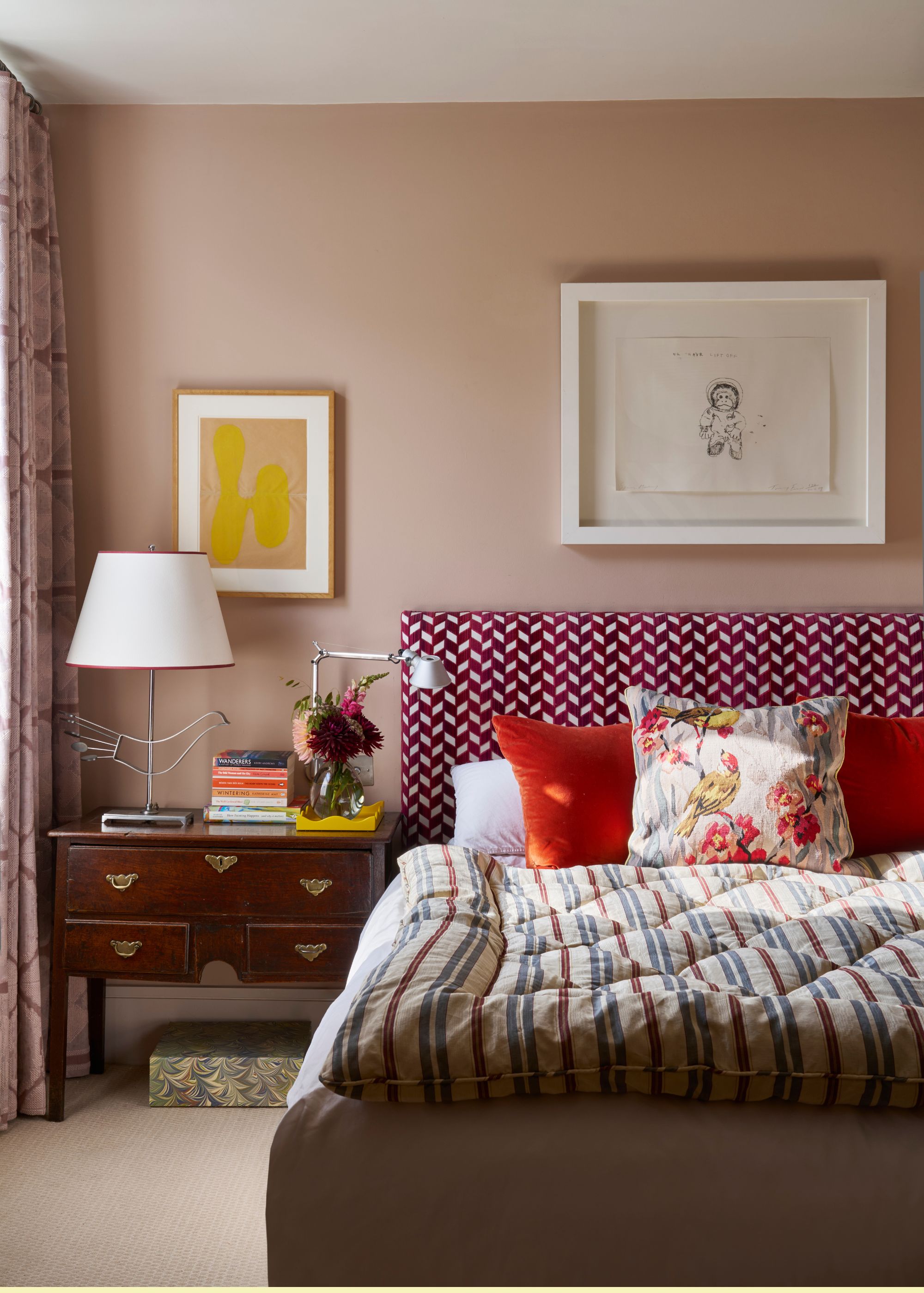 A double bed in a bedroom with a frame above the bed as well as a vintage beside table next to the bed. On the bedside table are stacked books, a table lamp and a reading lamp.