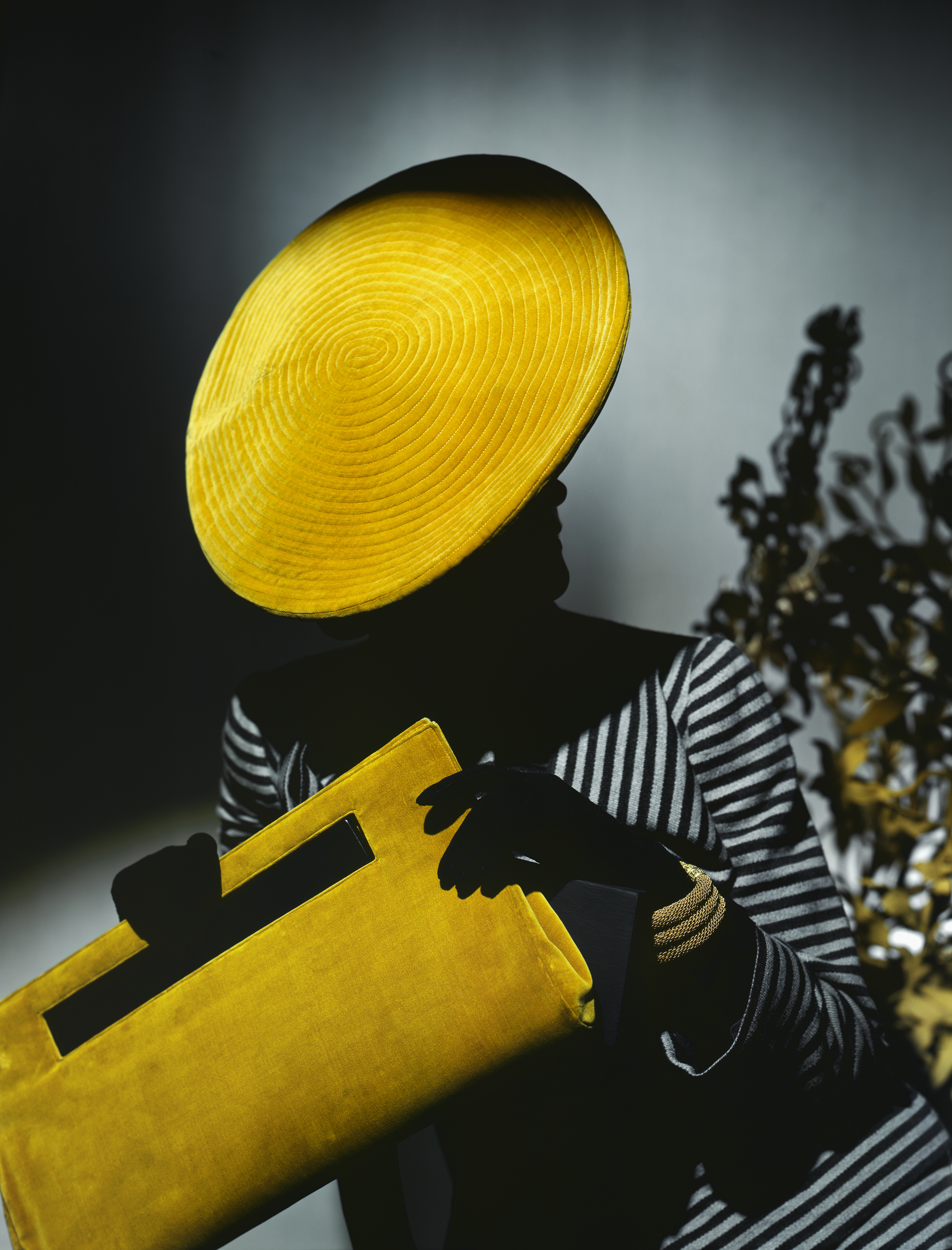 A stylized color photograph features a silhouette of a person wearing a bright yellow disc-shaped hat and holding a matching yellow clutch.