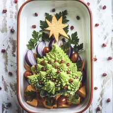 A baking tray decorated with a plant-based Christmas tree, made of Romanesco cauliflower and pomegranate arils, cherry tomatoes, red onion, sweet potato and carrots and garnished with Italian parsley. A white wooden, rustic background with a natural pattern of pomegranate seeds.