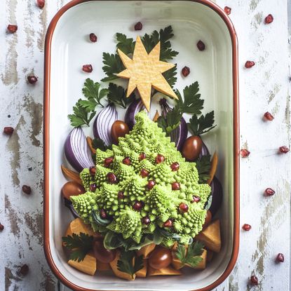 A baking tray decorated with a plant-based Christmas tree, made of Romanesco cauliflower and pomegranate arils, cherry tomatoes, red onion, sweet potato and carrots and garnished with Italian parsley. A white wooden, rustic background with a natural pattern of pomegranate seeds.