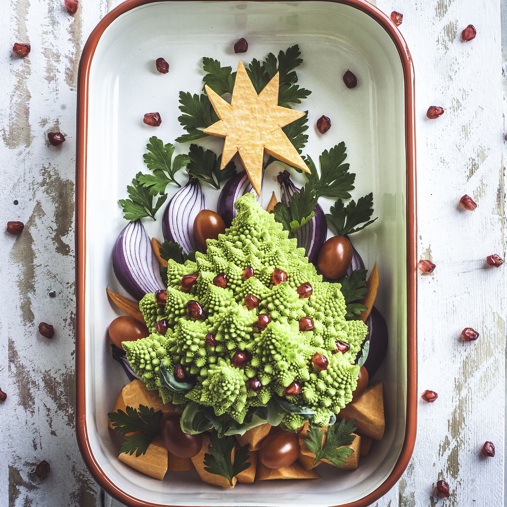 A baking tray decorated with a plant-based Christmas tree, made of Romanesco cauliflower and pomegranate arils, cherry tomatoes, red onion, sweet potato and carrots and garnished with Italian parsley. A white wooden, rustic background with a natural pattern of pomegranate seeds.