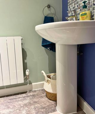 Side view of my sink, with a woven white basket with handles underneath. There is a dark green towel on a hoop on the wall and a pale green paint on the walls. There is a mosaic backsplash above the sink with a bottle of liquid Palmolive hand soap and chrome fixtures