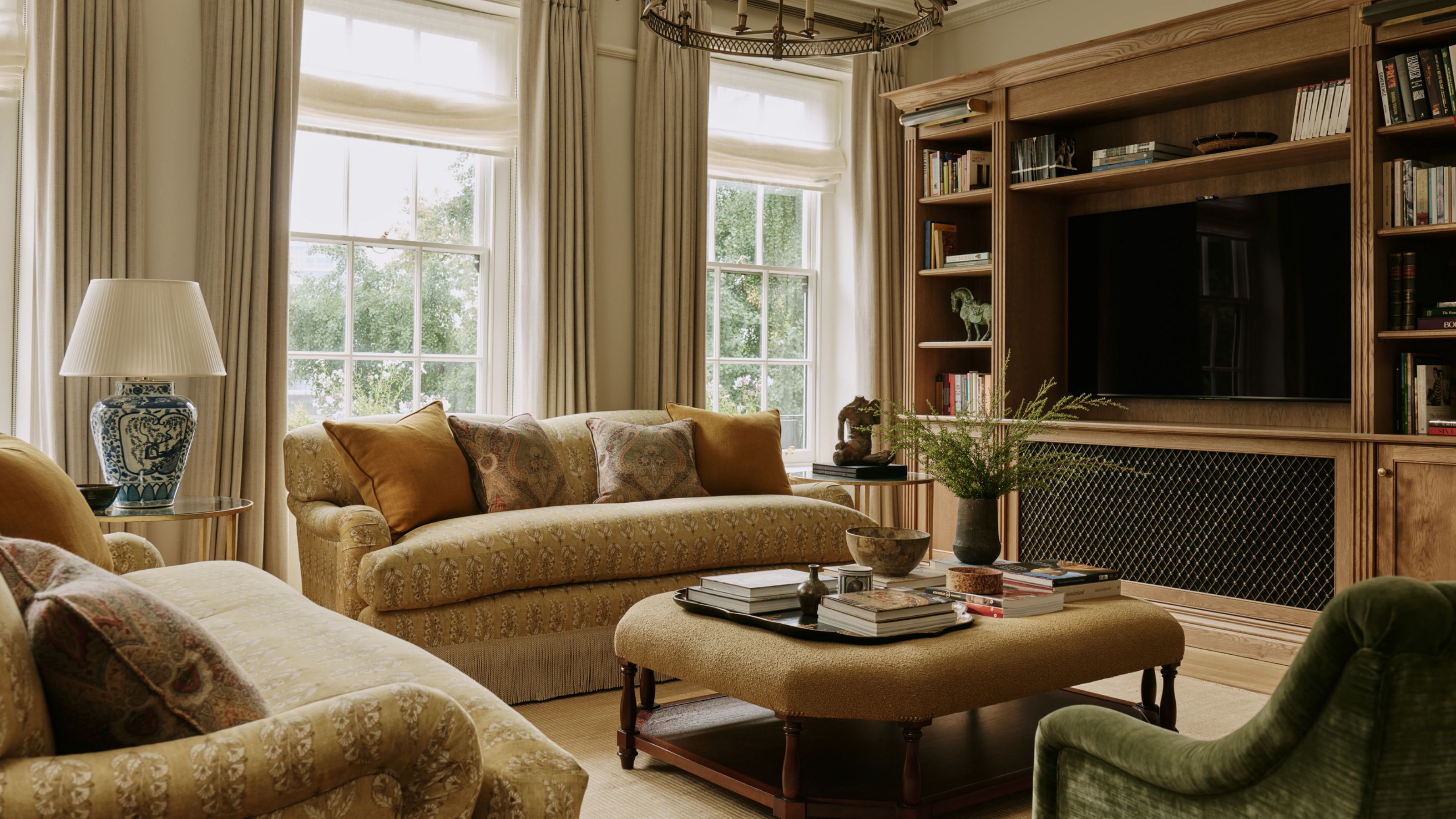a london living roo with large georgian windows, a large wooden media unit with built-in TV, two golden toned patterened sofas with fringe trim, ottoman and a green velvet chair
