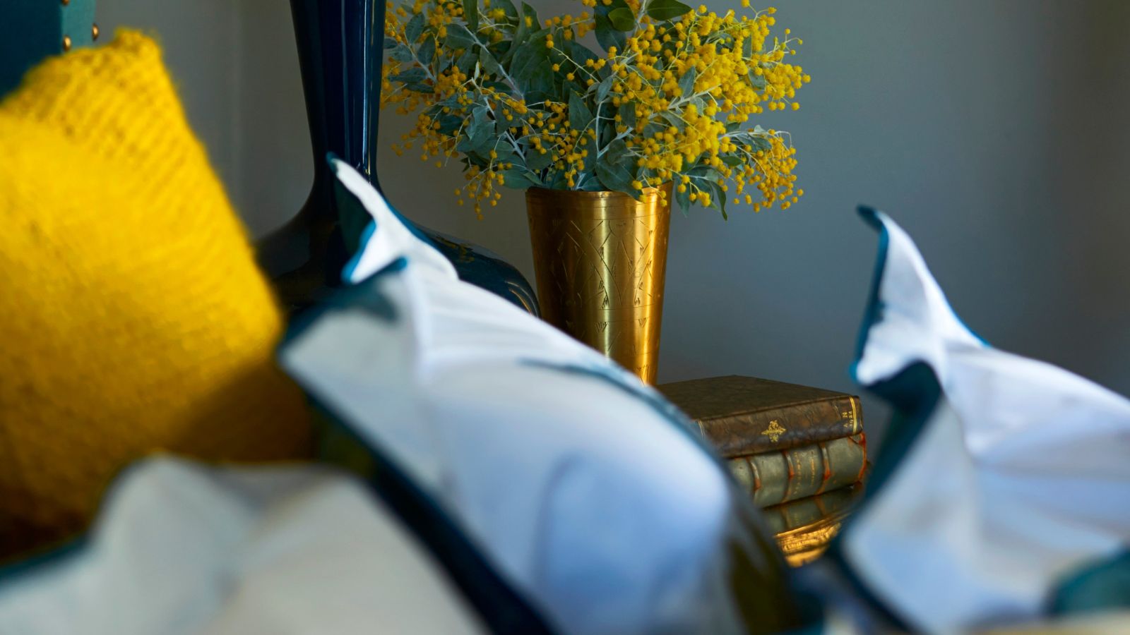 Blue bedroom with white pillows, gold cushions, and a gold vase filled with yellow flowers with green leaves.