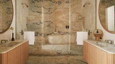stone-drenched bathroom with double floating timber-clad vanities on either side with marble counter and built-in sinks and round mirrors on the wall, and an enclosed shower with a shower seat and dual showerheads