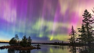 Northern lights erupt over a lake in Minnesota in a dark sky overhead shining rainbow of Aurora light as the sun sets on the horizon