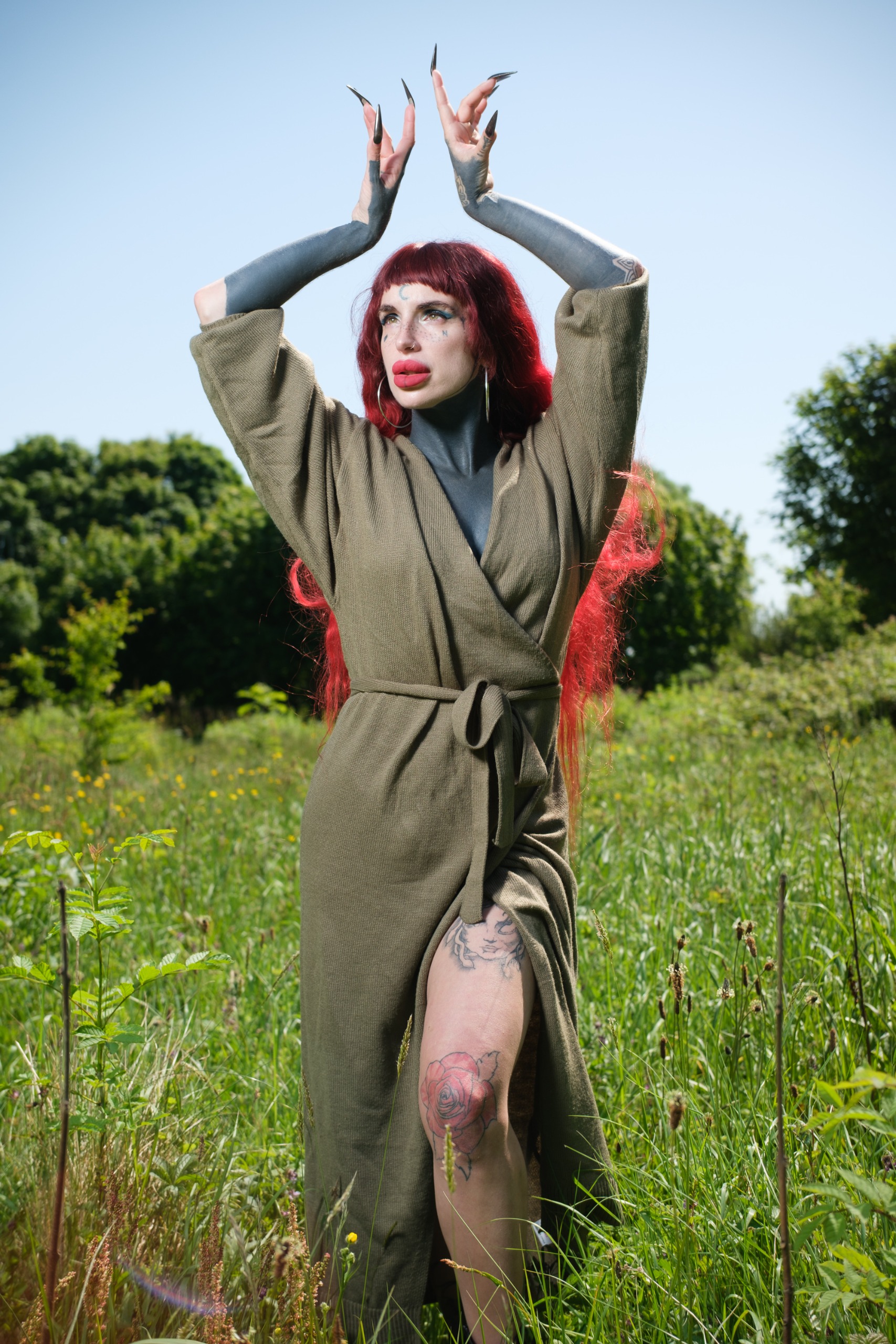 Close-up portrait of a red-haired person with dark body paint, hoop earrings, and bold makeup in front of a modern glass building.