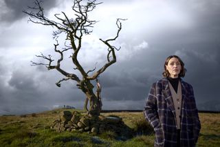 An image for Suranne Jones: Investigating Witch Trials showing Suranne Jones standing in a field. There is a small tree behind her, its branches twisted and gnarled, and there are no leaves on the tree. The sky behind her is full of dark clouds