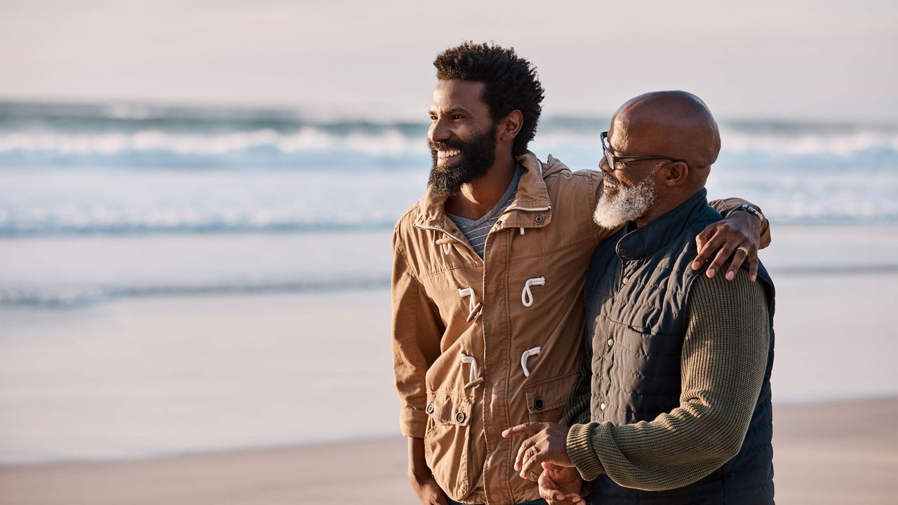 A young man walks with his gray-bearded dad on a beach