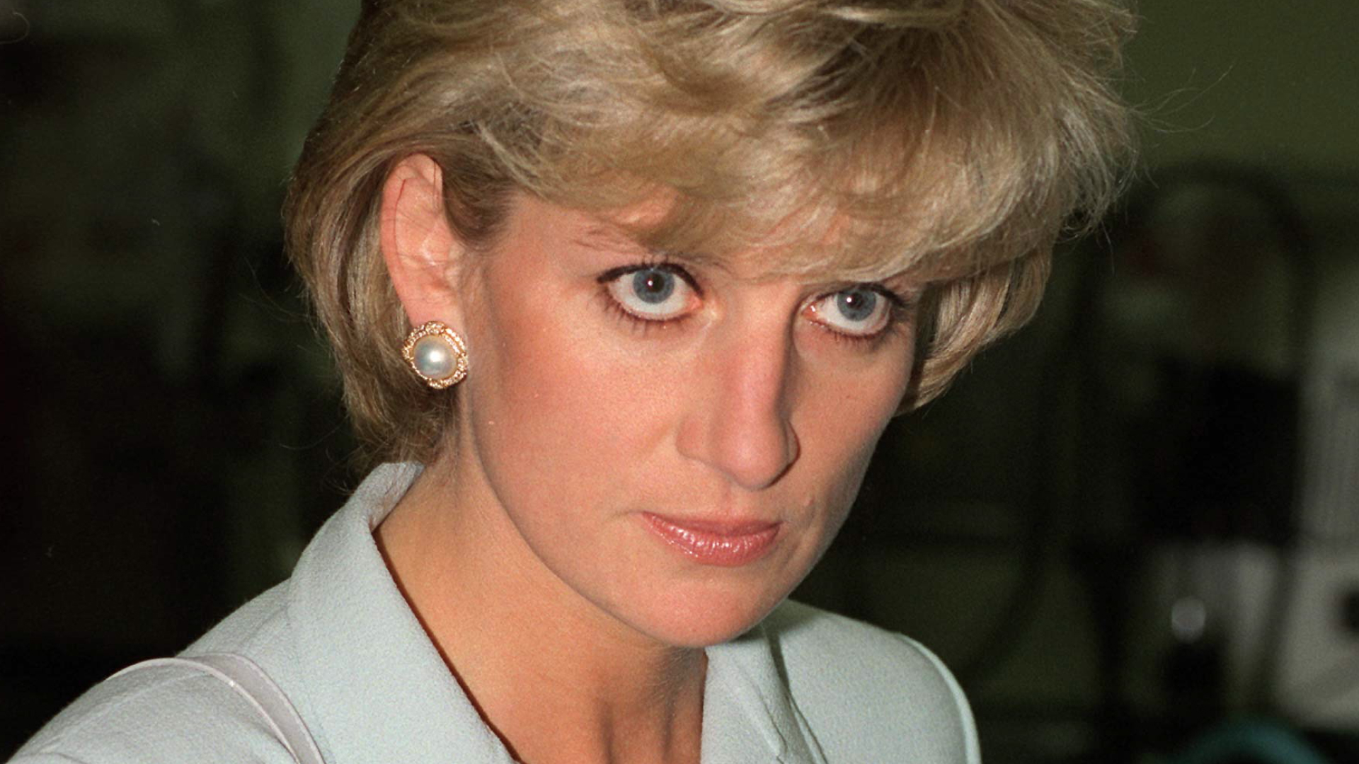 Princess Diana wearing a light blue skirt suit and large pearl earrings with a gold surround, she has short blonde hair