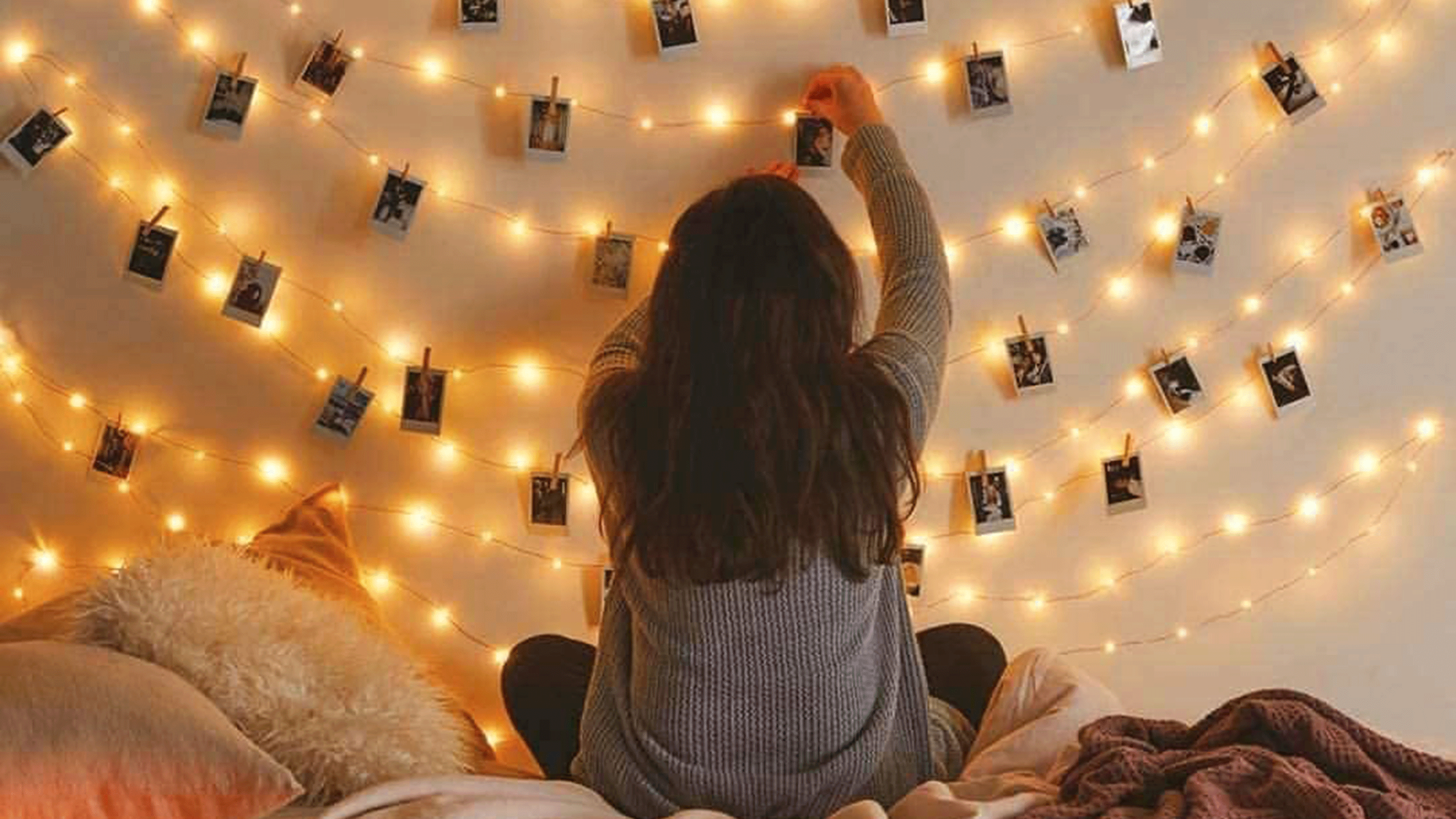 A female sitting on a bed fixing polaroid-style photographs onto bedroom fairy LED string lighting