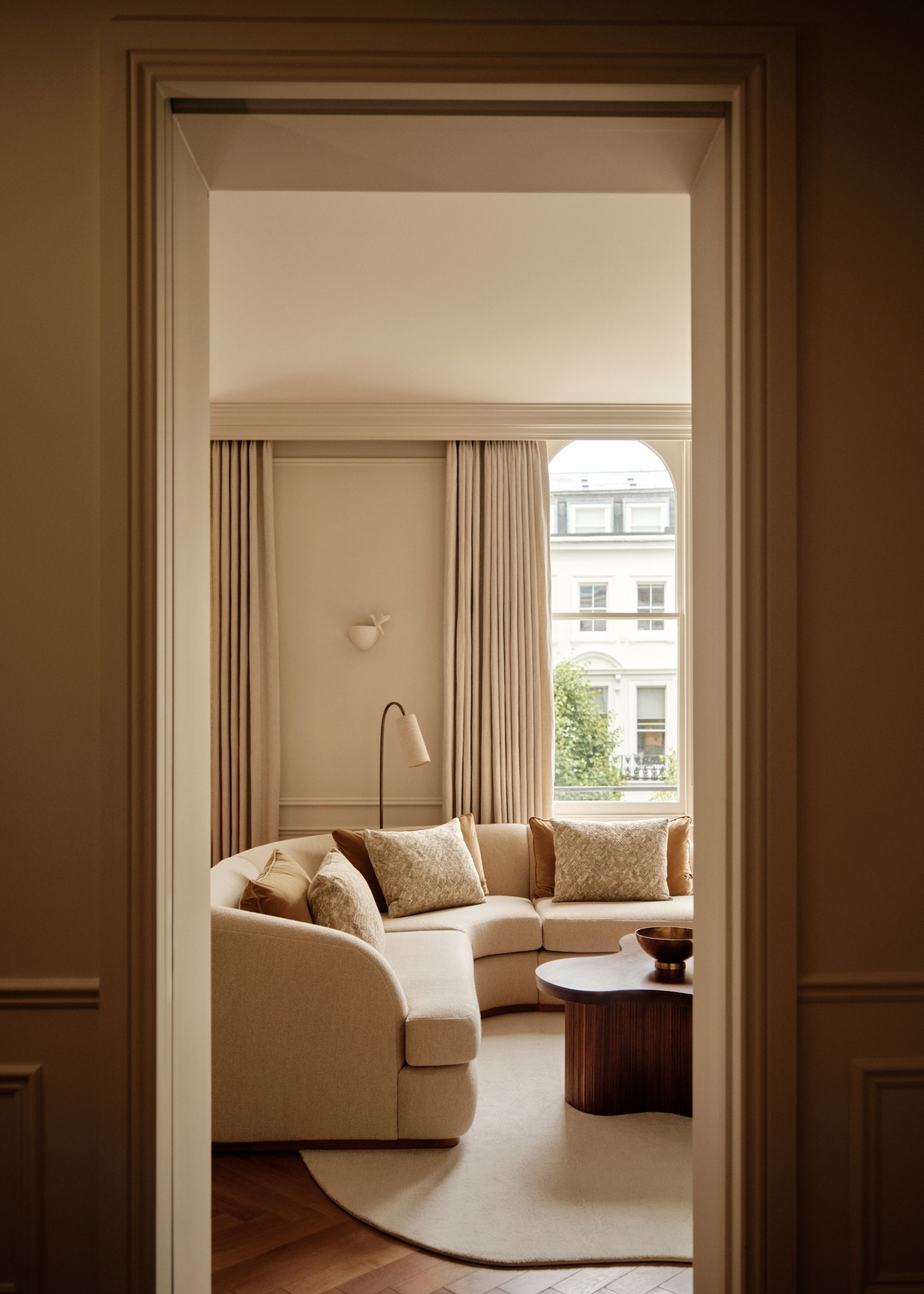 A doorway leading into a modern, neutral living room in a period home with soft neutral walls and curtains, an arched window, a large beige corner sofa, a dark wood sculptural coffee table and a cream rug.