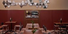 The interior of a retro-inspired trattoria features checkered floors, black leather chairs, wooden tables, and a reflective chrome wall decor, all imbued with a dark red and beige palette.
