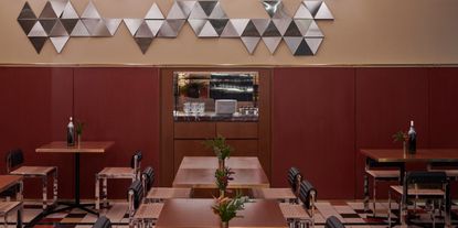 The interior of a retro-inspired trattoria features checkered floors, black leather chairs, wooden tables, and a reflective chrome wall decor, all imbued with a dark red and beige palette.