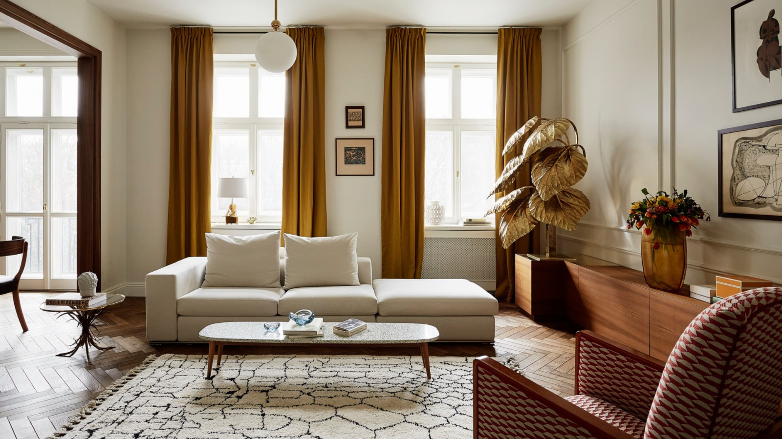 a modern living room with a white couch, mustard yellow curtains, a wooden sideboard, a black and white rug, and a gold-leafed plant