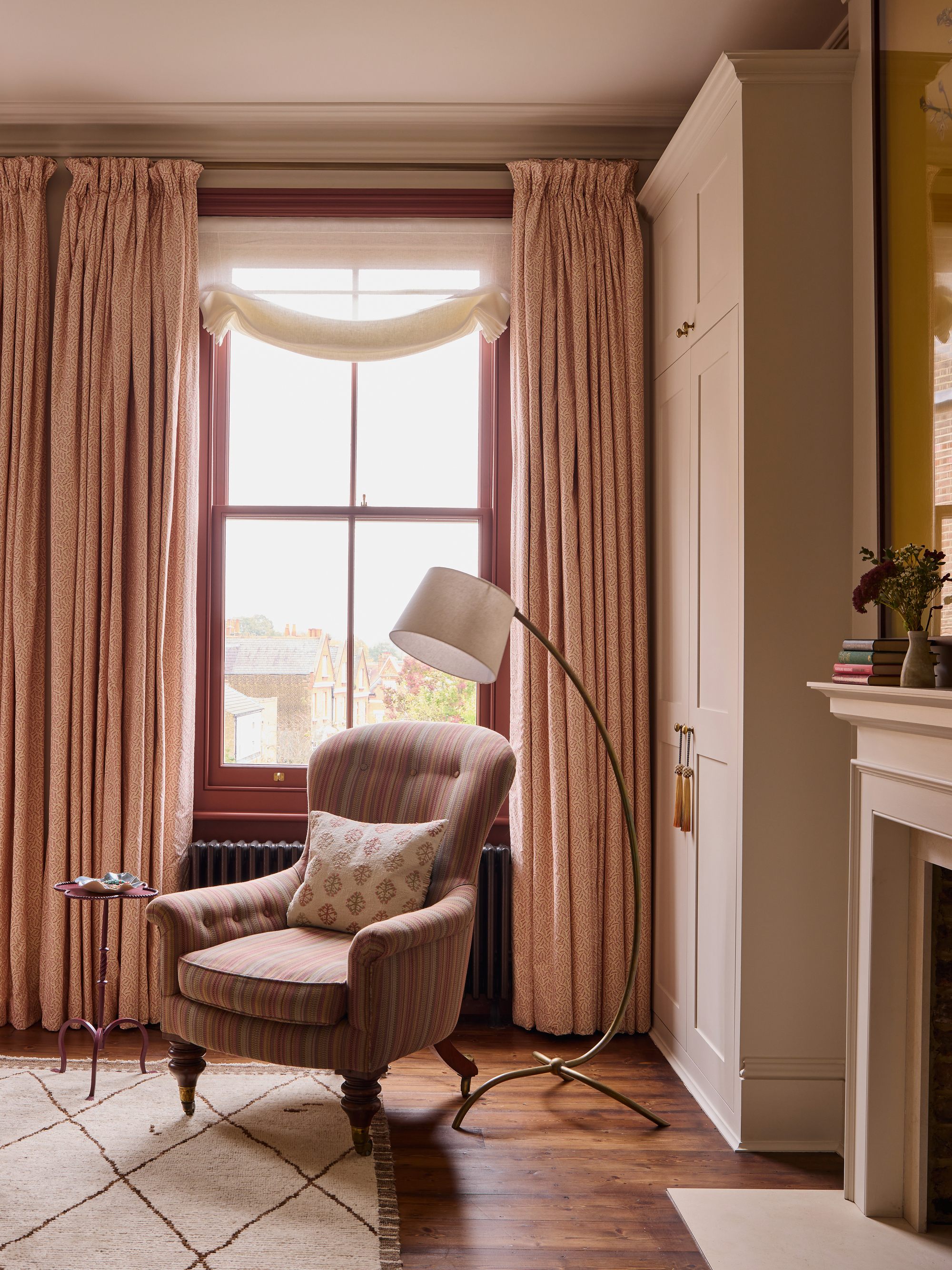 A reading corner in a room with beige walls and a beige wardrobe with a window that has a red painted window frame and pink curtains. There is a striped chair and a floor lamp.