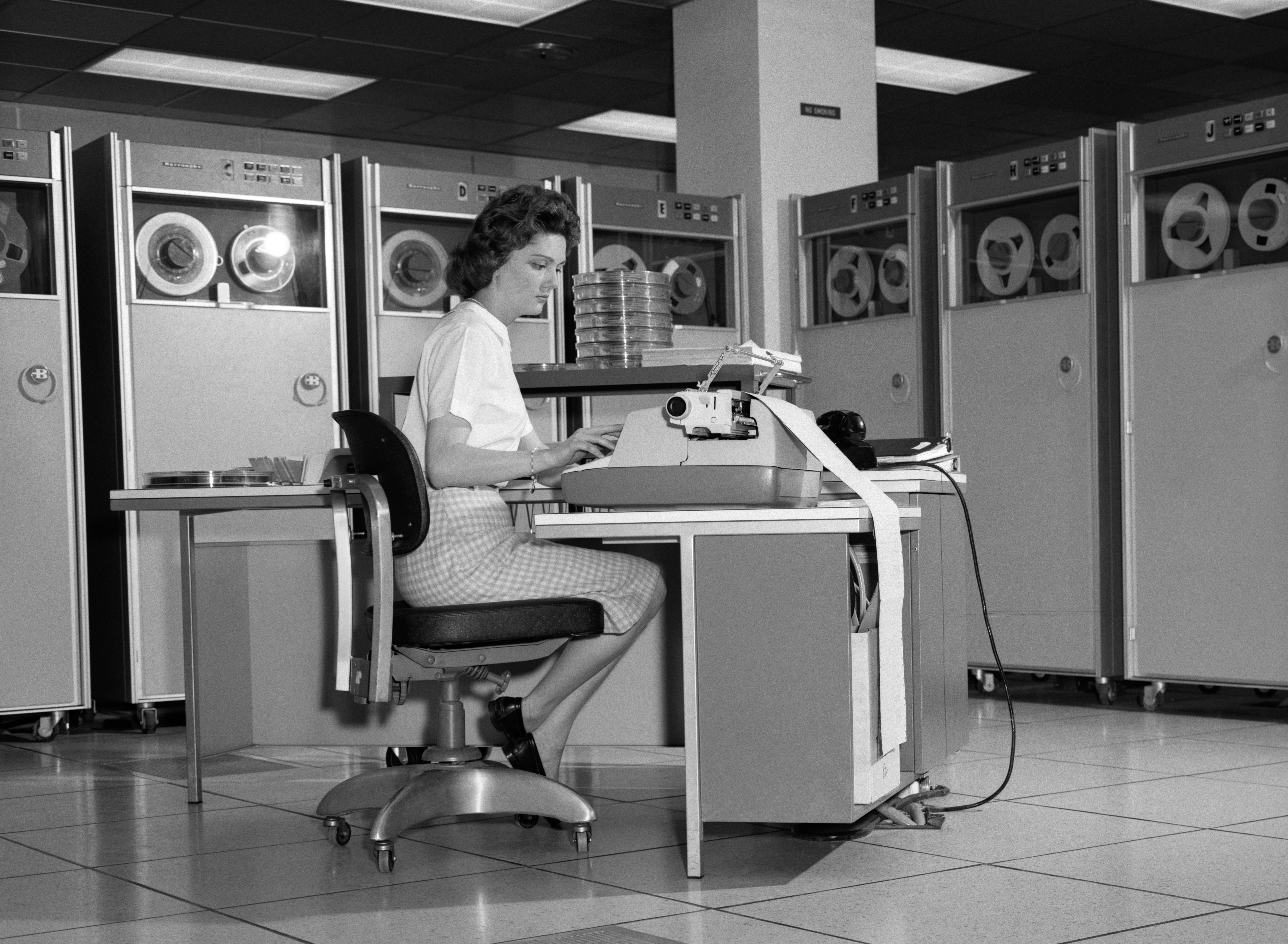 A black and white photo of a 1960s woman n a mainframe computer room, typing at typewriter