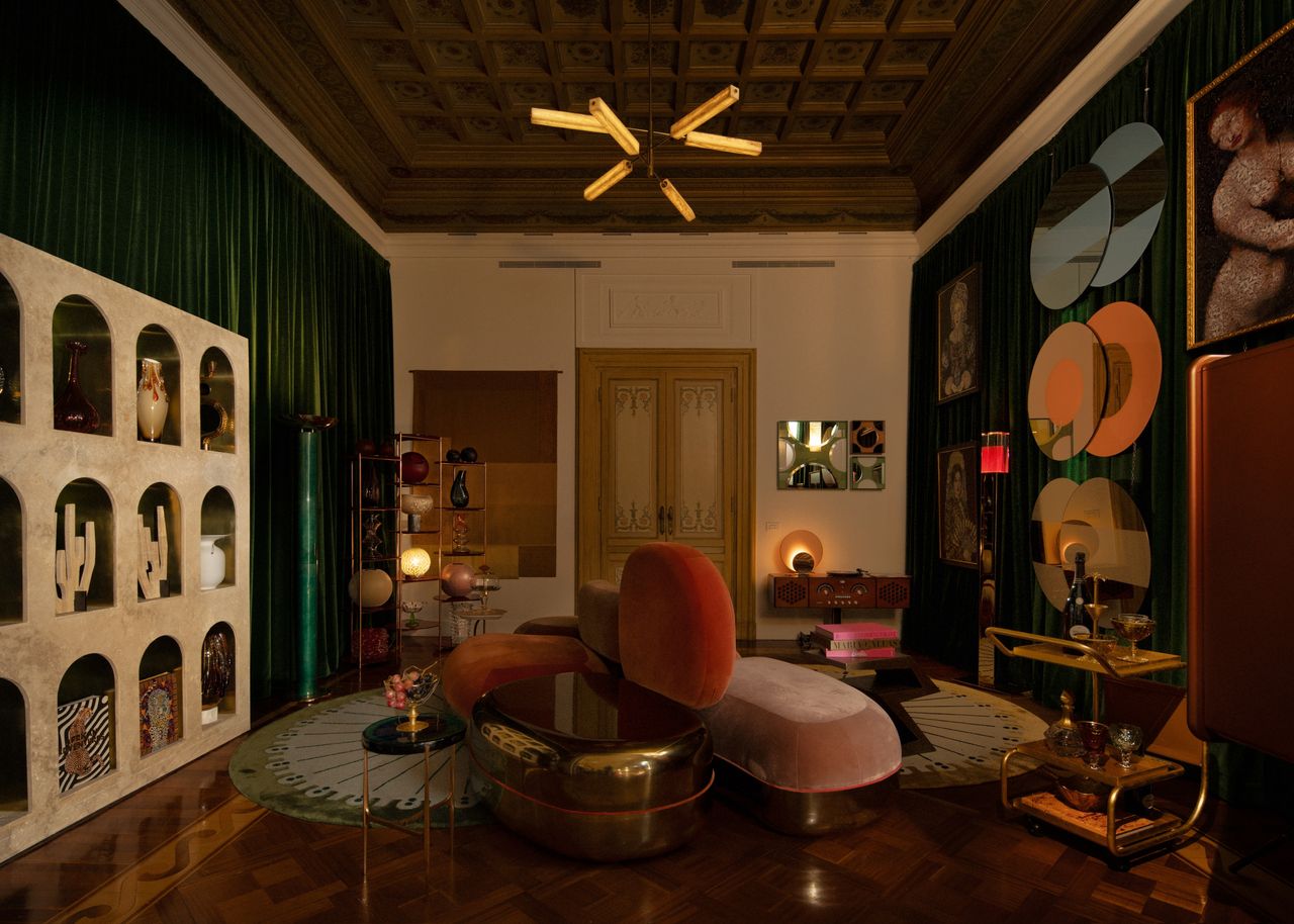 A moody concept space with circle artwork on the dark green-curtained wall with seating in the center on top of circular rugs with a dark paneled ceiling with a statement ceiling light, a bar cart, arched storage cubby holes and decor pieces all around