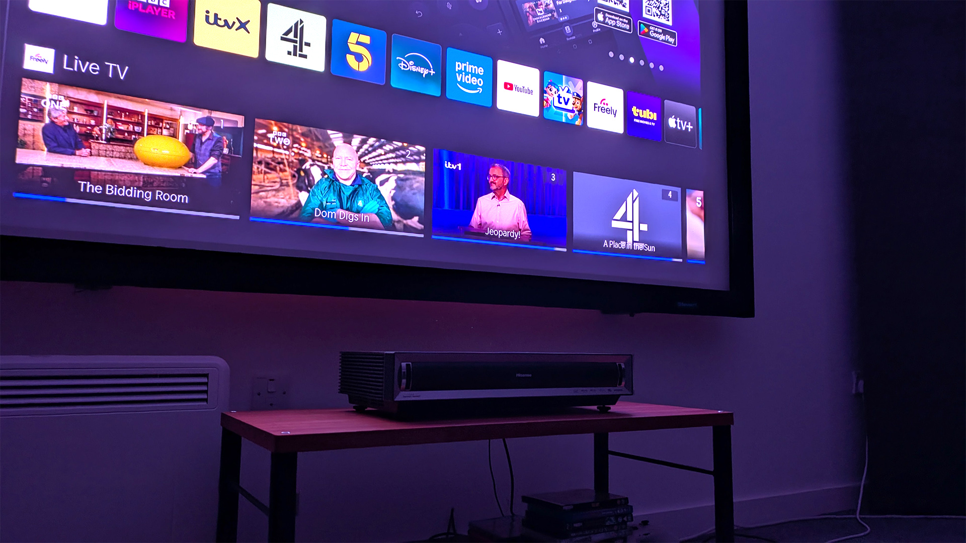 Hisense PX3 ultra short throw projector on wooden AV rack below TV showing Vidaa home screen